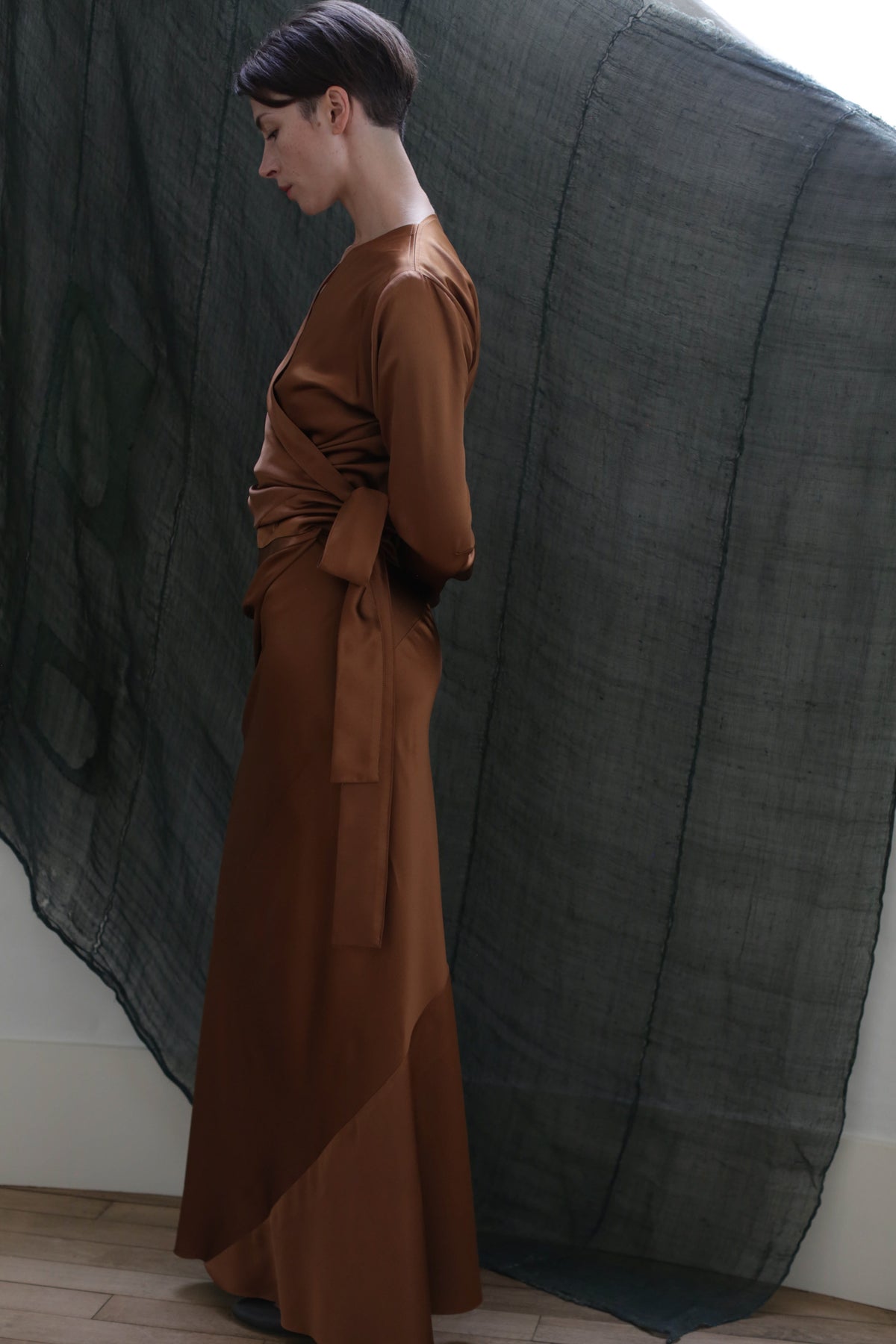 A woman with short dark hair stands in profile, wearing the X Blouse in Silk Charmeuse Copper, featuring a tied waist and elegant drape, against a sheer dark green fabric backdrop.