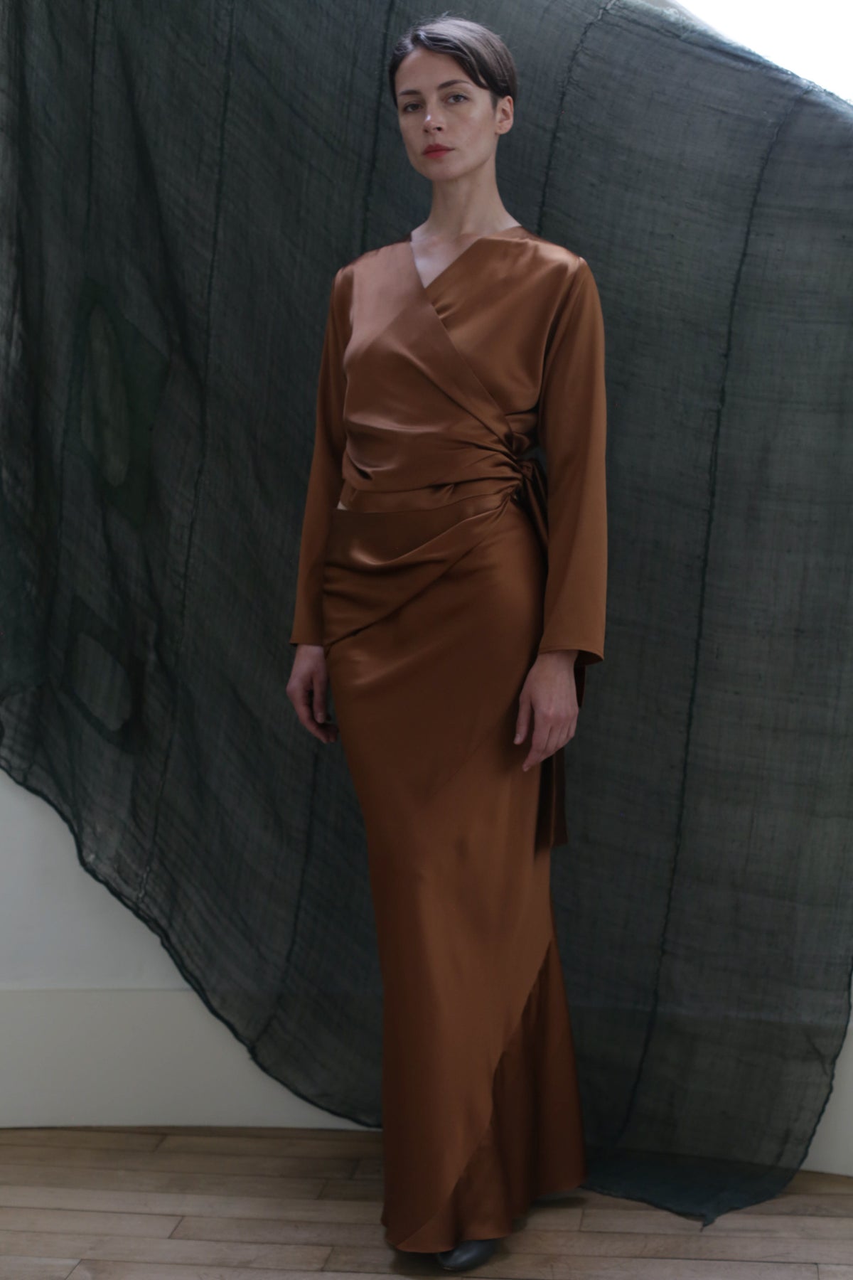 A woman stands before a sheer, dark textured backdrop wearing the Atelier Lan Jaenicke Dragon Skirt in silky copper Silk Charmeuse, featuring gathered waist detailing and a wrap-style bodice.