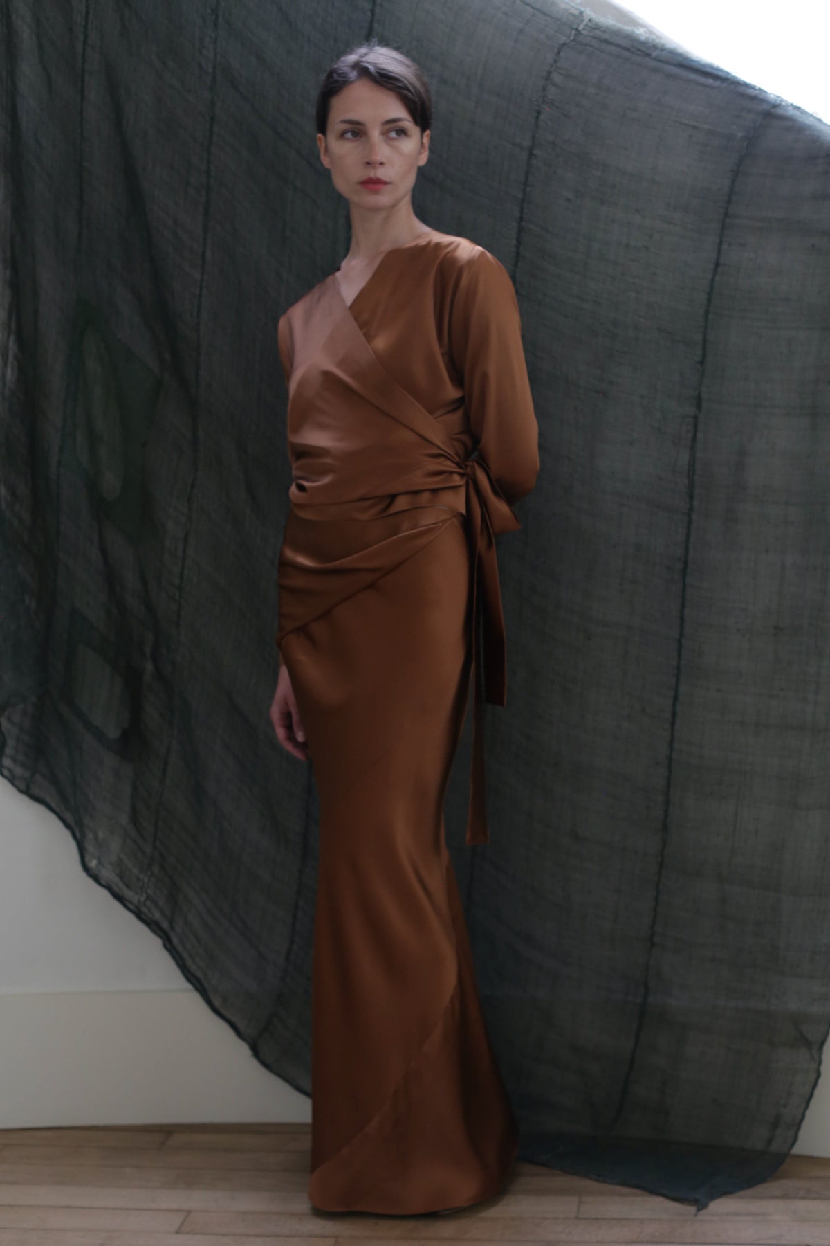 A woman wearing the Dragon Skirt in Silk Charmeuse Copper by Atelier Lan Jaenicke stands before a dark, sheer fabric backdrop, looking slightly to the side with a neutral expression.