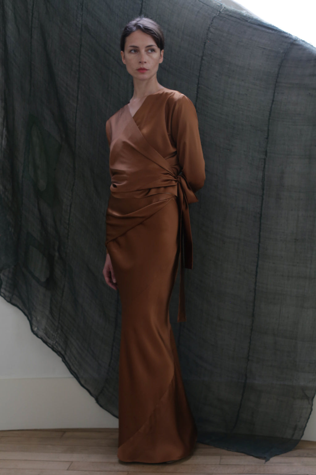 A woman stands before a sheer, dark backdrop, wearing the X Blouse in Silk Charmeuse Copper. She faces slightly to the side, gazing calmly at the camera.