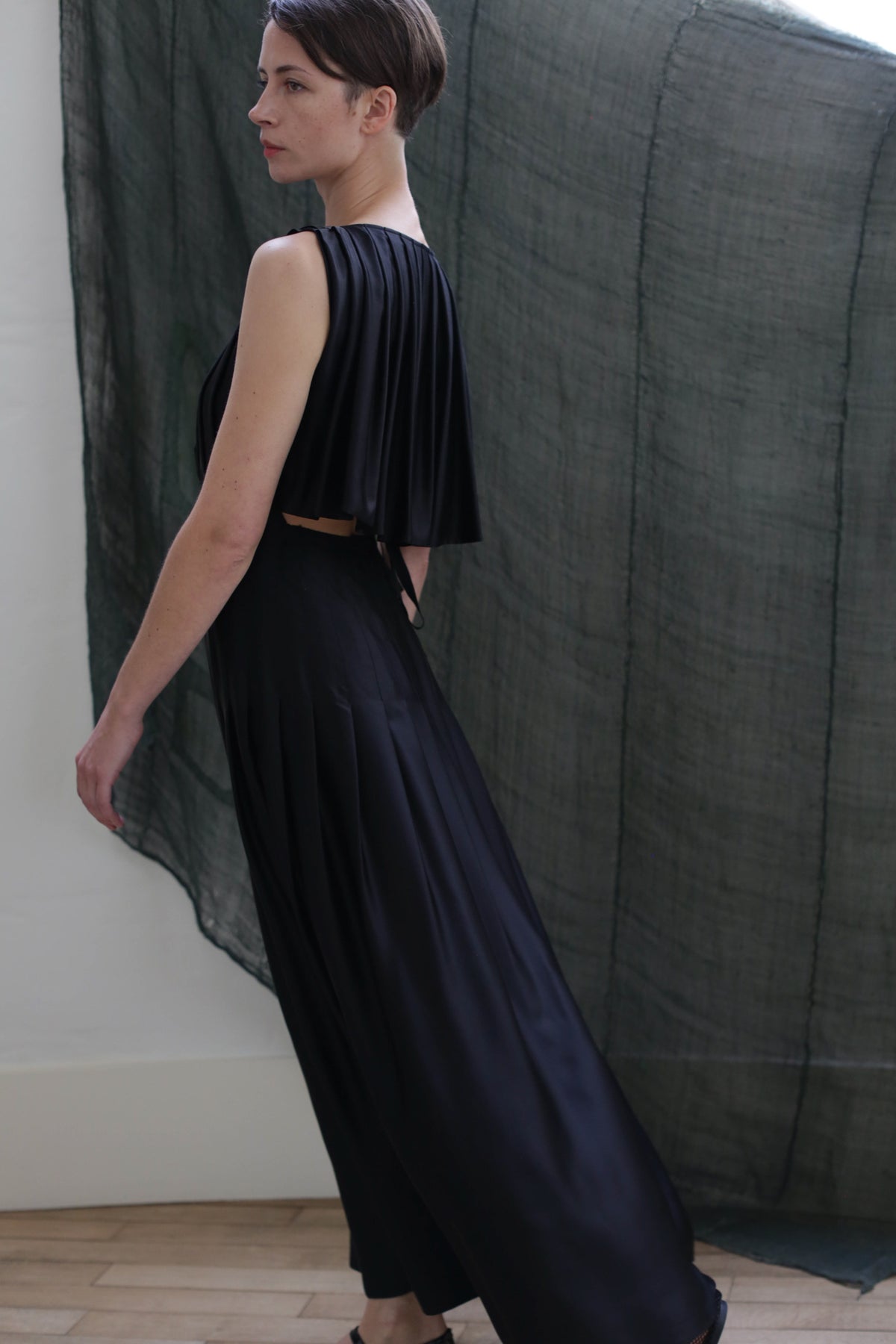 A person with short hair models the Hand Pleated Column Dress in Silk Charmeuse Black, a sleeveless, floor-length pleated gown. Standing sideways against draped dark sheer fabric, the handmade design creates an elegant, sophisticated silhouette.