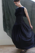 A person wears the Hand Pleated Column Dress in black silk charmeuse, posing lightly turned to the side while holding the skirt. Sheer dark fabric drapes on the wall behind her above a light-colored floor.