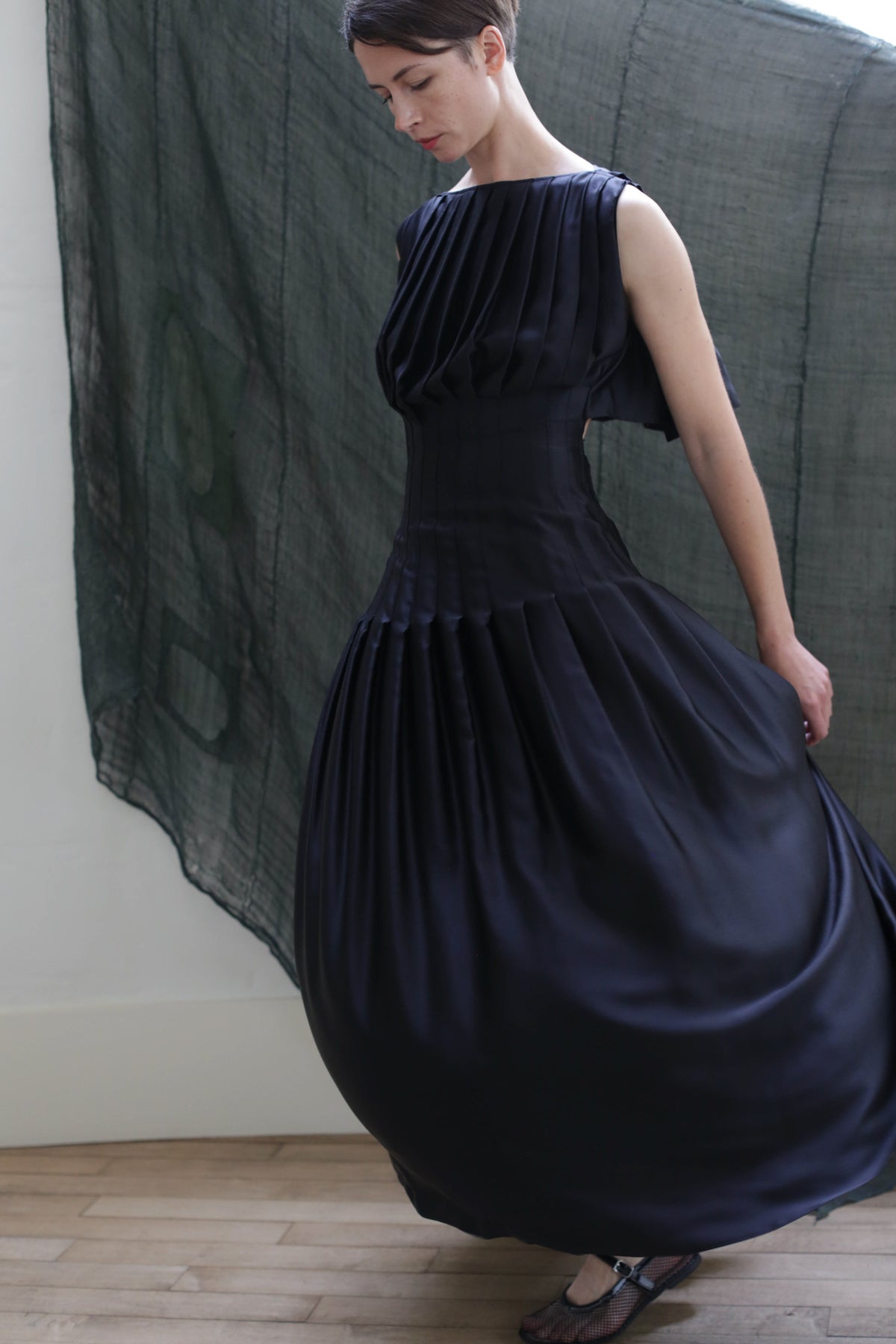 A person wears the Hand Pleated Column Dress in black silk charmeuse, posing lightly turned to the side while holding the skirt. Sheer dark fabric drapes on the wall behind her above a light-colored floor.