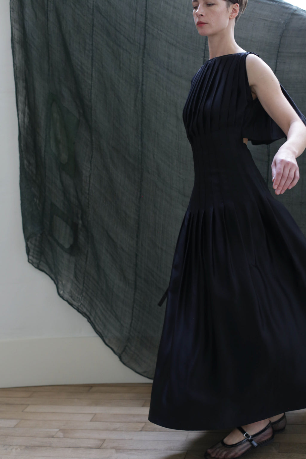 A woman wears the Hand Pleated Column Dress in Silk Charmeuse Black, posing with one arm extended before a sheer dark green backdrop on a wooden floor. Her face is partially out of frame.