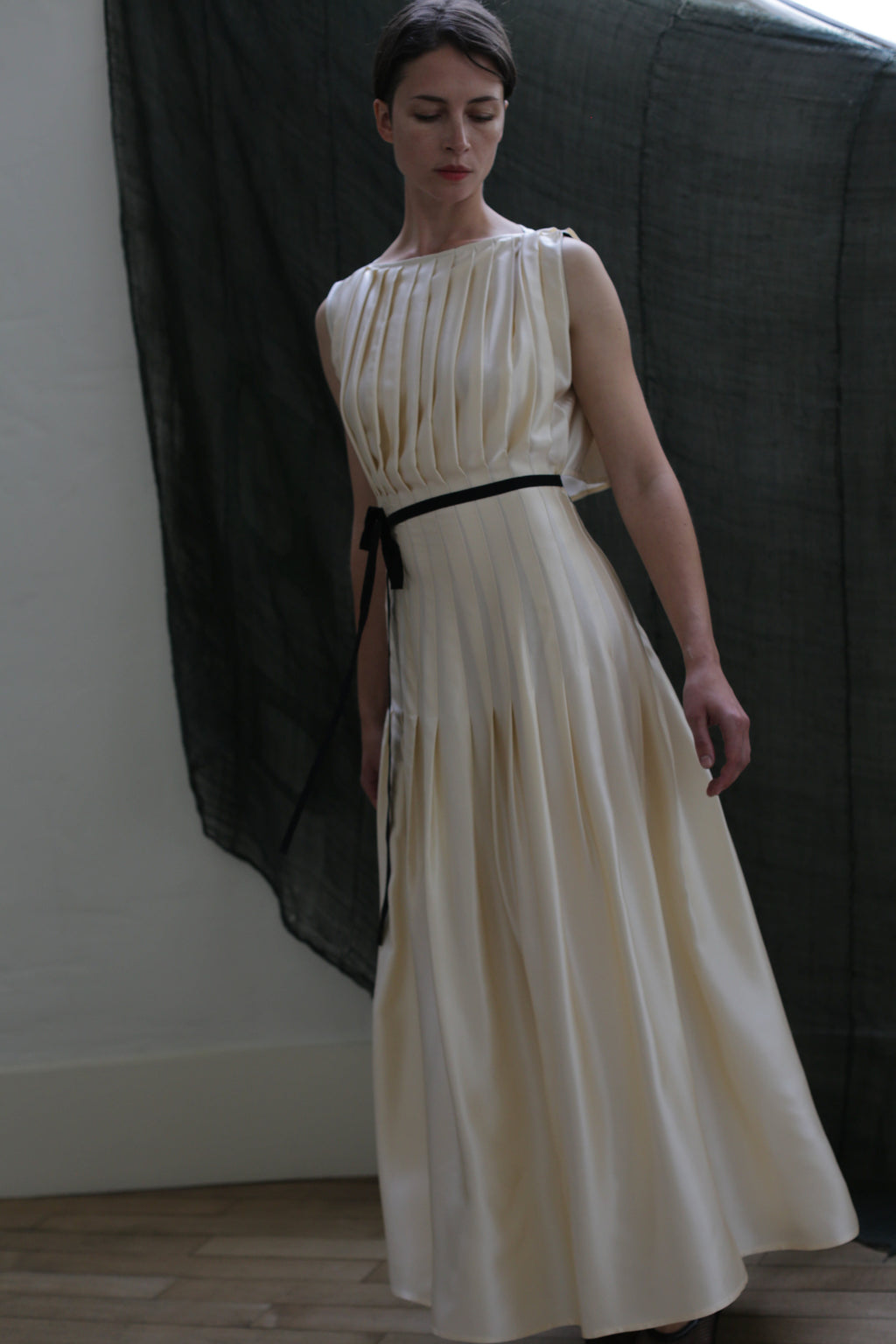 Wearing the Hand Pleated Column Dress in silk charmeuse ivory from Atelier Lan Jaenicke, a person with a black ribbon belt stands before a dark sheer curtain, looking down with a neutral expression.