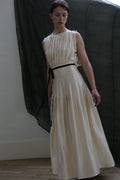 Wearing the Hand Pleated Column Dress in silk charmeuse ivory from Atelier Lan Jaenicke, a person with a black ribbon belt stands before a dark sheer curtain, looking down with a neutral expression.