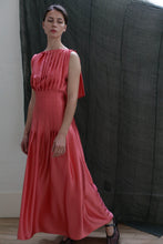 Load image into Gallery viewer, A woman stands indoors before a dark textured backdrop, wearing the Hand Pleated Column Dress in silk charmeuse sakura—a sleeveless, floor-length Grecian-inspired gown with hand-stitched pleats—paired with black heels.