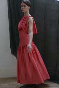 A woman poses gracefully indoors on a wooden floor, wearing the Hand Pleated Column Dress in Silk Charmeuse Sakura, with one arm extended and a dark, sheer fabric hanging in the background.