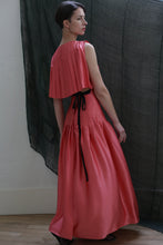 Load image into Gallery viewer, A woman in the Hand Pleated Column Dress | Silk Charmeuse Sakura, featuring hand-stitched pleats and a black ribbon tie, stands near a sheer black curtain, gazing over her shoulder.