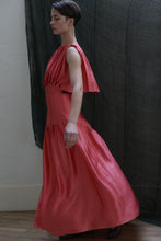 Load image into Gallery viewer, A person in profile wears the Hand Pleated Column Dress | Silk Charmeuse Sakura, a flowing sleeveless coral dress with hand-stitched pleats and a cape-like shoulder detail, set against a neutral backdrop with dark fabric behind.