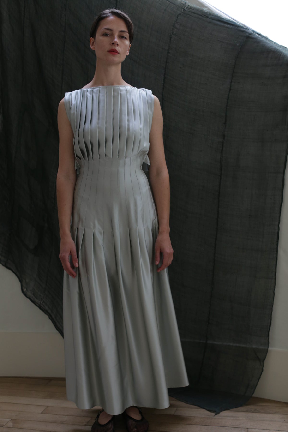 A woman stands before a sheer dark backdrop, wearing a sleeveless Hand Pleated Column Dress in Silk Charmeuse Celadon with ankle-length pleats. She faces forward, arms relaxed at her sides.