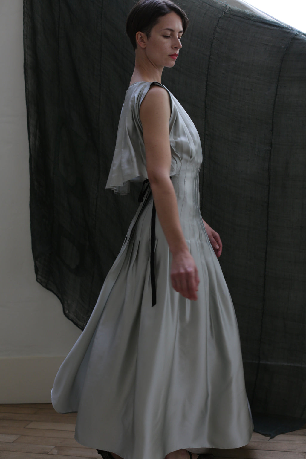 A person in profile wears the Hand Pleated Column Dress in Silk Charmeuse Celadon, its elegant fit and draped, hand-pleated details highlighted against a dark sheer background by soft, natural light.
