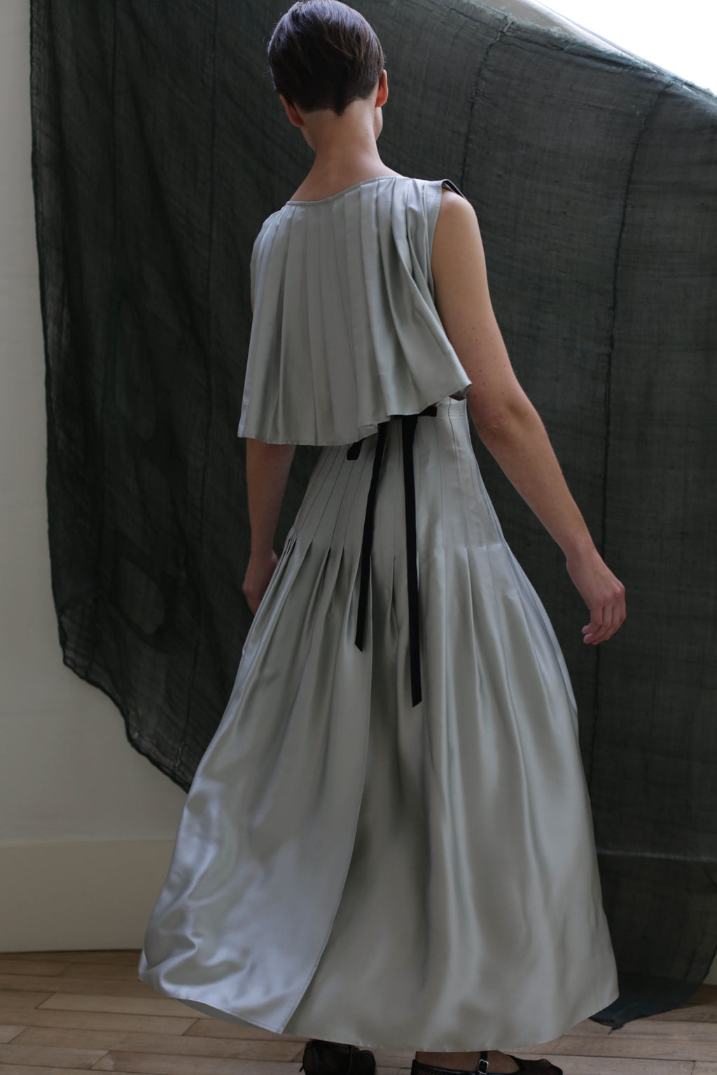 A person with short hair stands with their back to the camera, wearing the Hand Pleated Column Dress in silk charmeuse celadon, featuring light gray pleats and black ribbon details, in front of a dark sheer curtain and white wall.