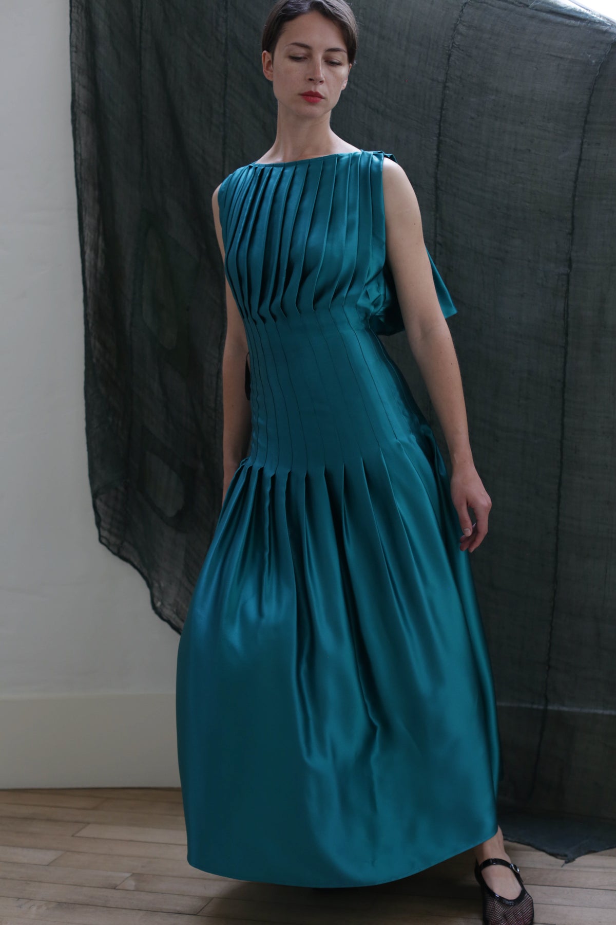 A woman in a sleeveless, floor-length teal Hand Pleated Column Dress | Silk Charmeuse Kiji stands indoors against a dark sheer backdrop, showcasing the elegant craftsmanship of Handmade Atelier Lan Jaenicke.