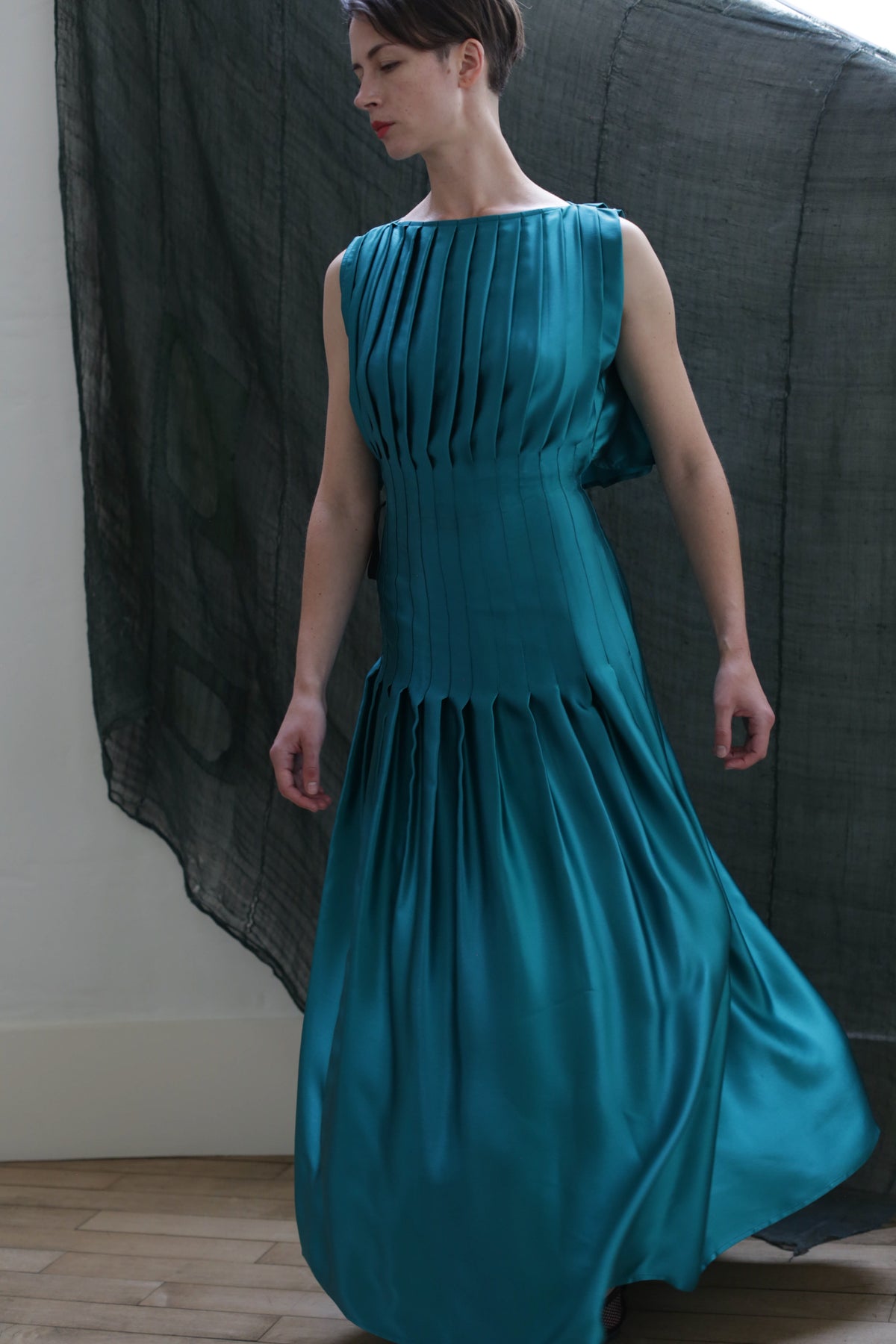 A person in the Hand Pleated Column Dress | Silk Charmeuse Kiji stands indoors before a dark, sheer hanging fabric, wooden floorboards visible beneath their feet.