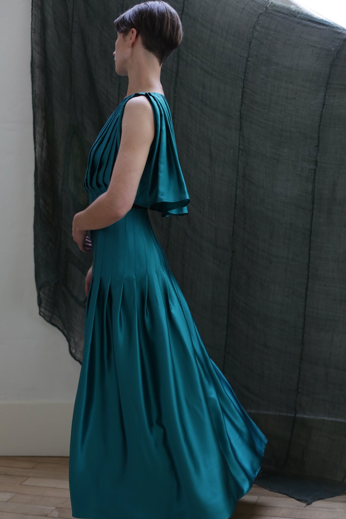 A person with short hair in profile wears the Hand Pleated Column Dress | Silk Charmeuse Kiji, standing indoors against a sheer dark fabric backdrop.