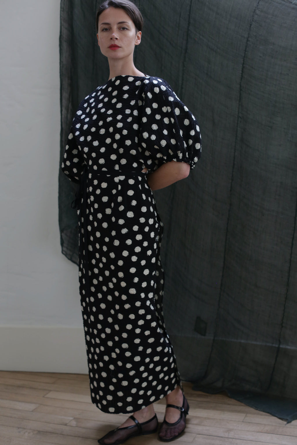 A woman stands indoors wearing the Kimono Dress | Polkadot Seersucker Black, featuring white polka dots and short puff sleeves. Her hands are behind her back, with a dark sheer curtain in the background.