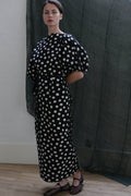 A woman stands indoors wearing the Kimono Dress | Polkadot Seersucker Black, featuring white polka dots and short puff sleeves. Her hands are behind her back, with a dark sheer curtain in the background.