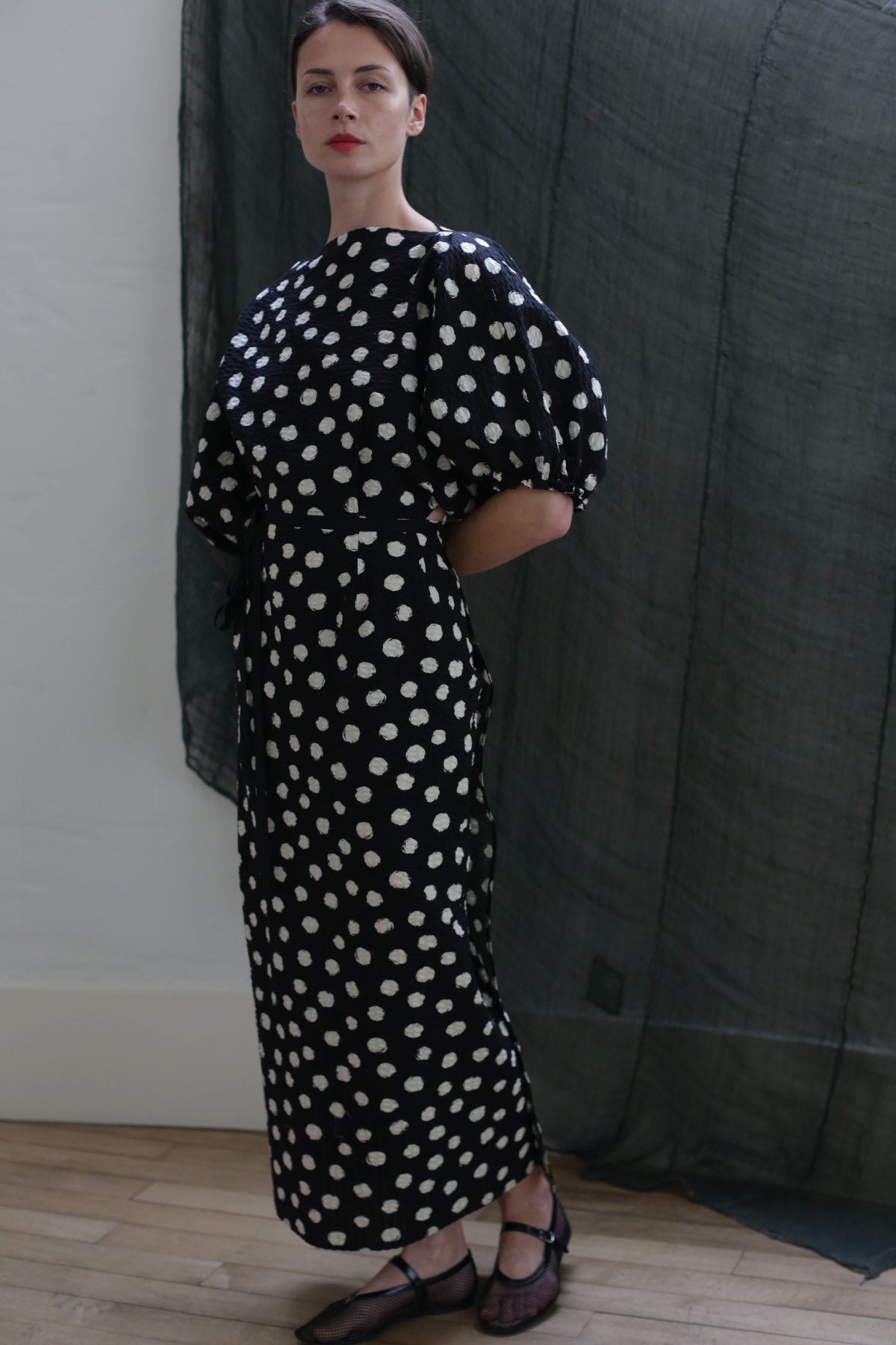 A woman stands indoors wearing the Kimono Dress | Polkadot Seersucker Black, featuring white polka dots and short puff sleeves. Her hands are behind her back, with a dark sheer curtain in the background.