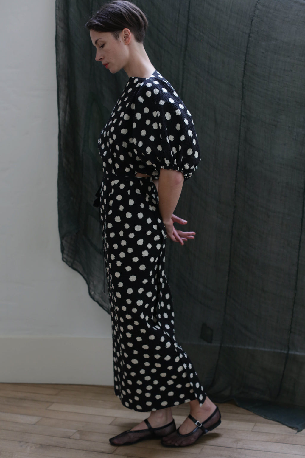A person stands sideways indoors wearing the Kimono Dress | Polkadot Seersucker Black, featuring black polka-dot fabric and subtle balloon sleeves. Their hands are clasped behind their back, with dark sheer fabric hanging in the background.