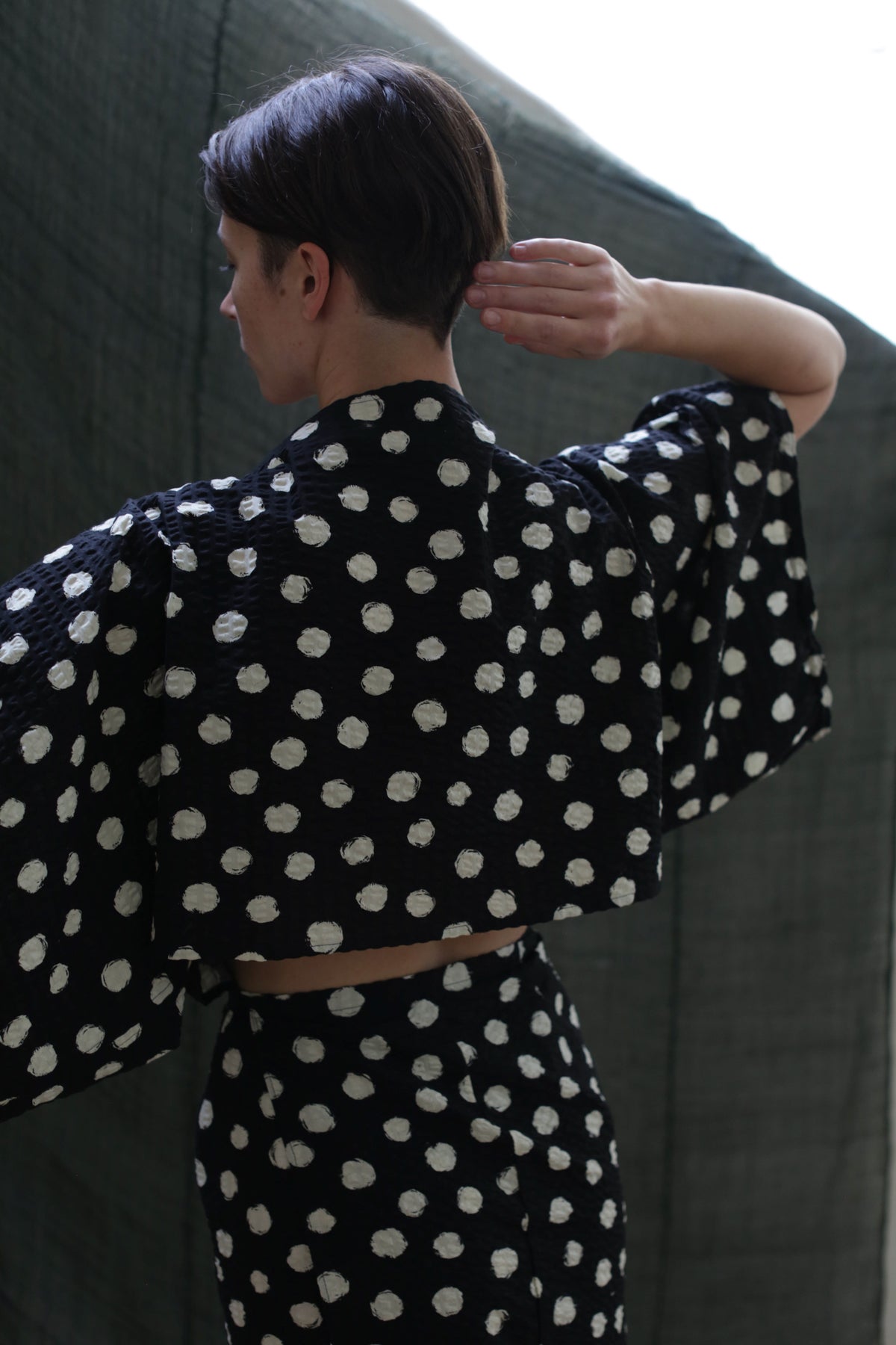 A person with short dark hair, seen from behind against a dark green background, wears the Kimono Dress | Polkadot Seersucker Black, featuring white polka dots and wide sleeves as they touch their neck.