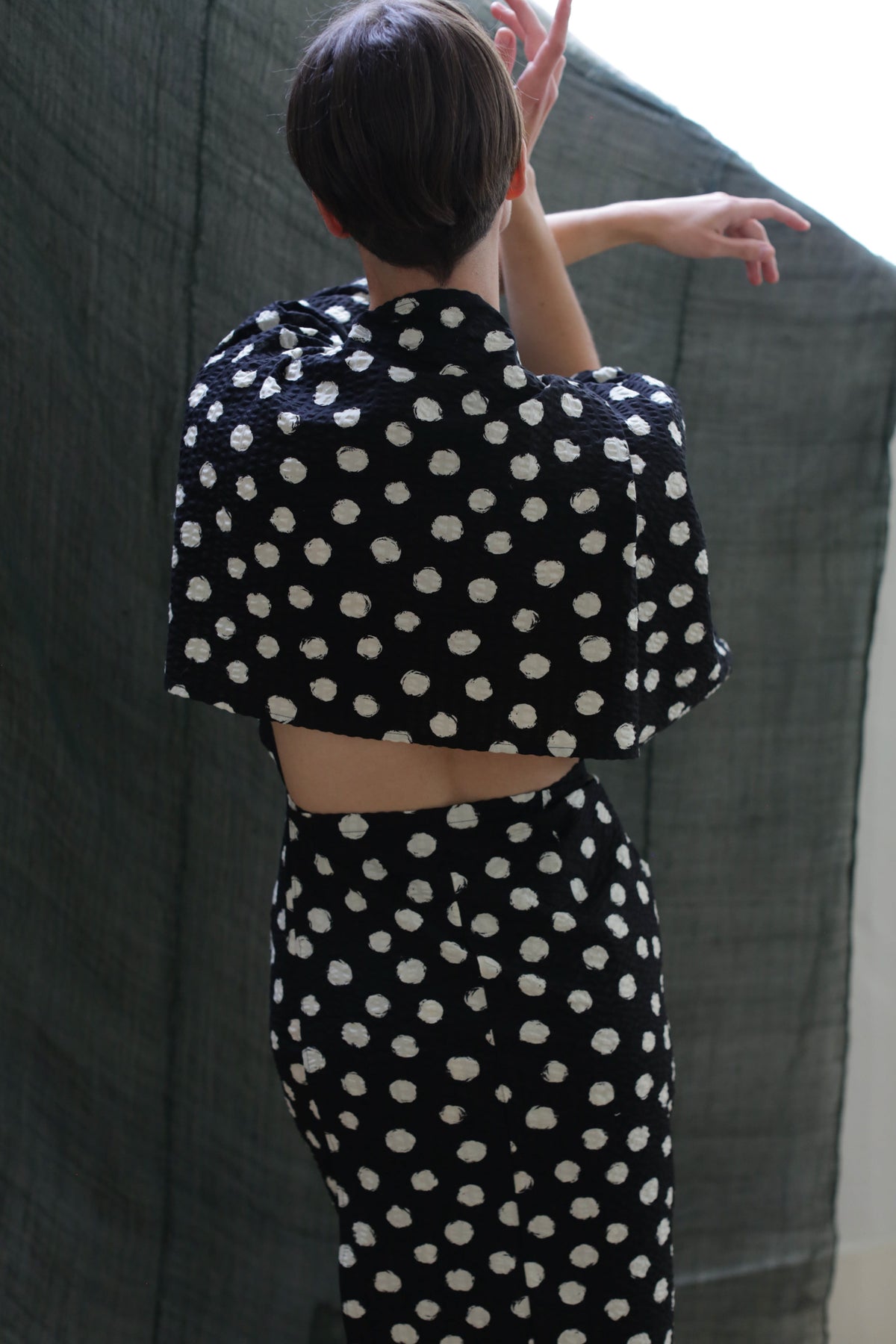 A person with short hair, arms raised and back to the camera, wears the Kimono Dress | Polkadot Seersucker Black, featuring large white polka dots and an open back. A dark, textured fabric forms the background.