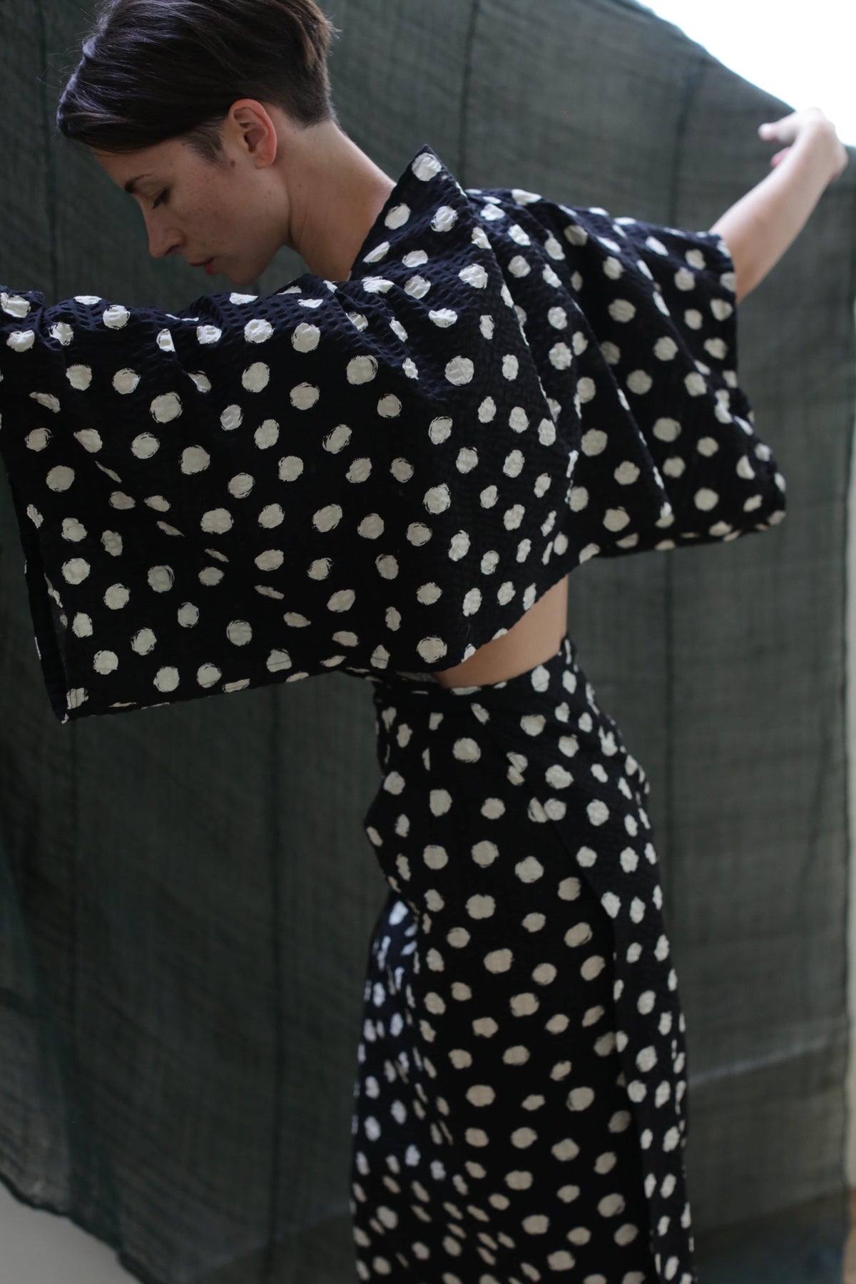 A person models the Kimono Dress | Polkadot Seersucker Black, featuring large white polka dots, wide sleeves, and an open back, standing sideways with arms raised against a dark fabric backdrop.
