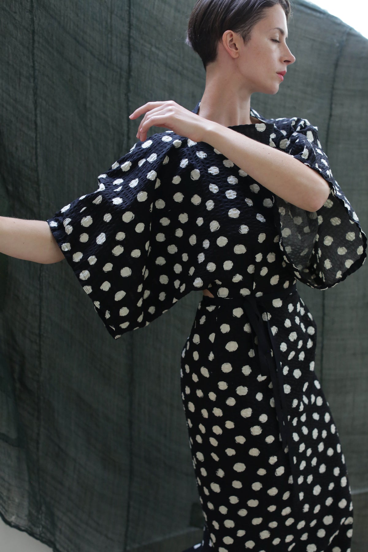 A person with short brown hair poses in the Kimono Dress | Polkadot Seersucker Black, featuring wide sleeves and a loose fit, against a dark green fabric backdrop.