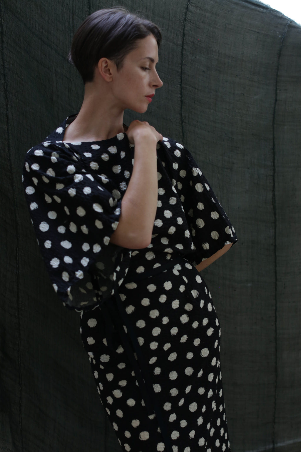 A woman with short brown hair wears the Kimono Dress | Polkadot Seersucker Black—featuring black fabric with large white polka dots and balloon sleeves—as she stands before a dark green textured backdrop, looking down thoughtfully.