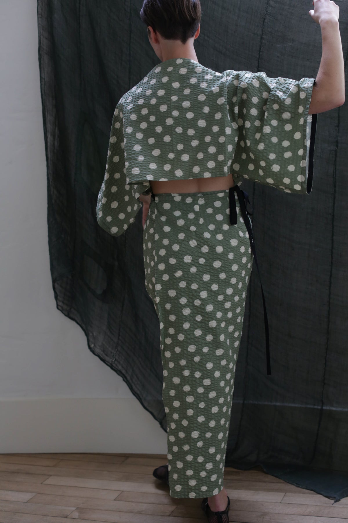 A person stands with their back to the camera, wearing the Kimono Dress | Polkadot Seersucker Moss featuring wide sleeves and a matching high-waisted skirt. One arm is raised, touching dark sheer fabric beside them, highlighting its Kimono-inspired design.