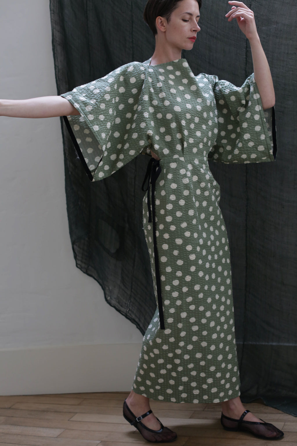 A person poses gracefully with arms raised, wearing the Kimono Dress | Polkadot Seersucker Moss—featuring a green polkadot pattern, kimono-inspired design, and wide sleeves—in front of a sheer dark curtain.