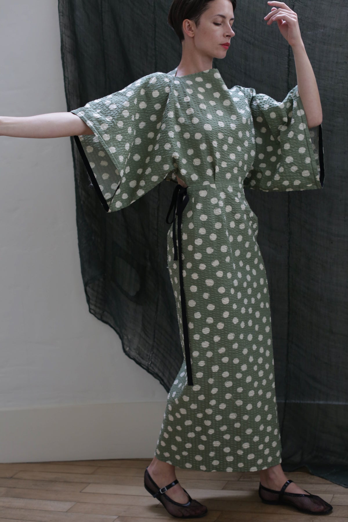 A person poses gracefully with arms raised, wearing the Kimono Dress | Polkadot Seersucker Moss—featuring a green polkadot pattern, kimono-inspired design, and wide sleeves—in front of a sheer dark curtain.