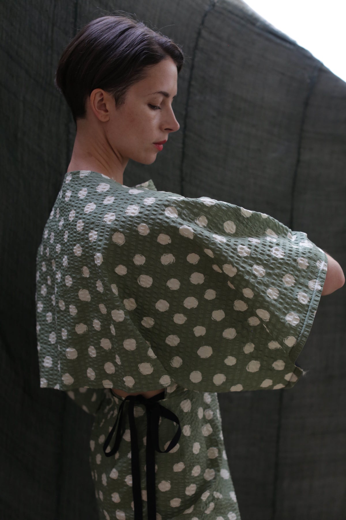 A woman with short brown hair stands in profile, wearing the Kimono Dress | Polkadot Seersucker Moss with wide sleeves, set against a dark textured background.
