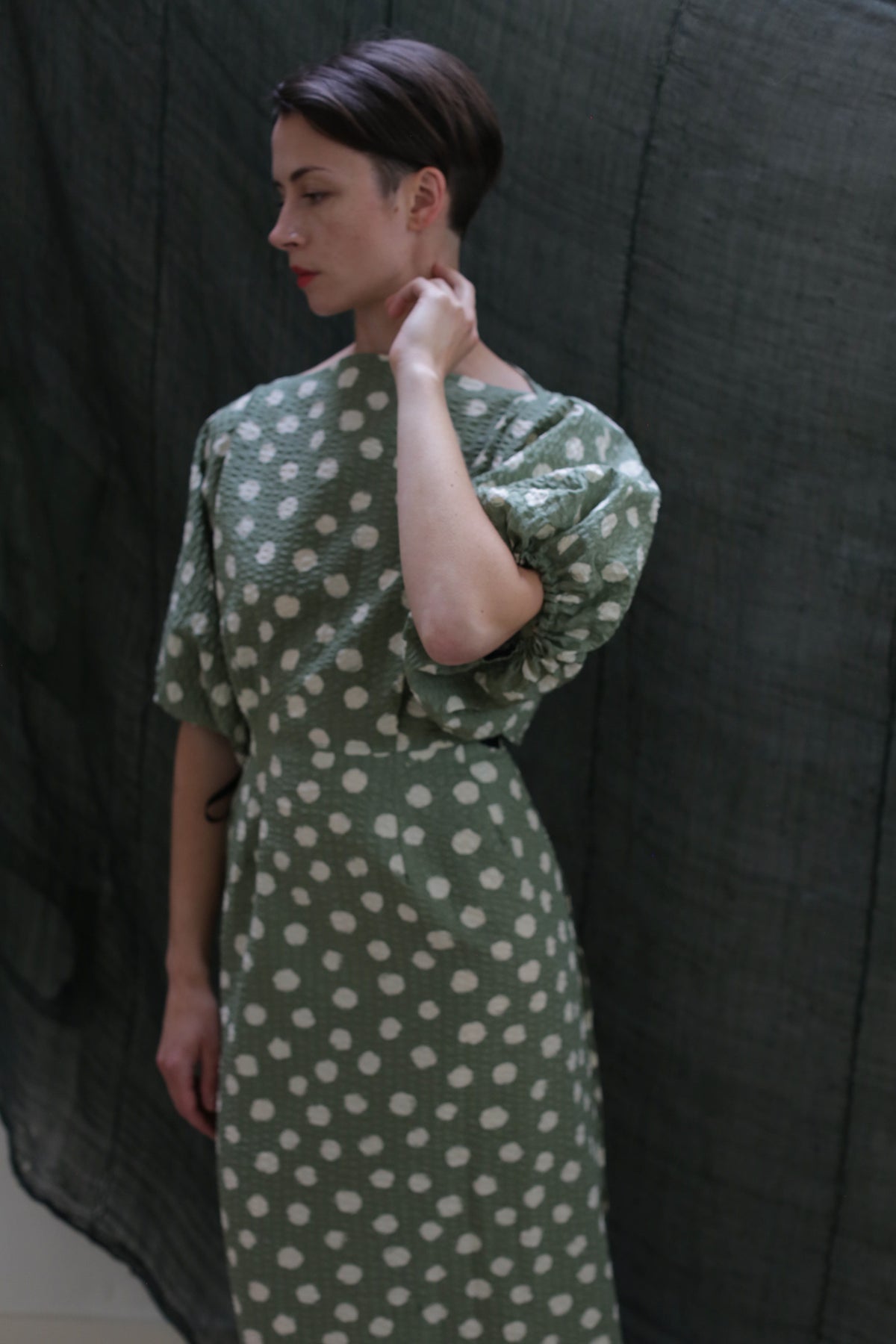 A person wearing the Kimono Dress | Polkadot Seersucker Moss stands before a dark textured background, gently touching their neck and gazing to the side.