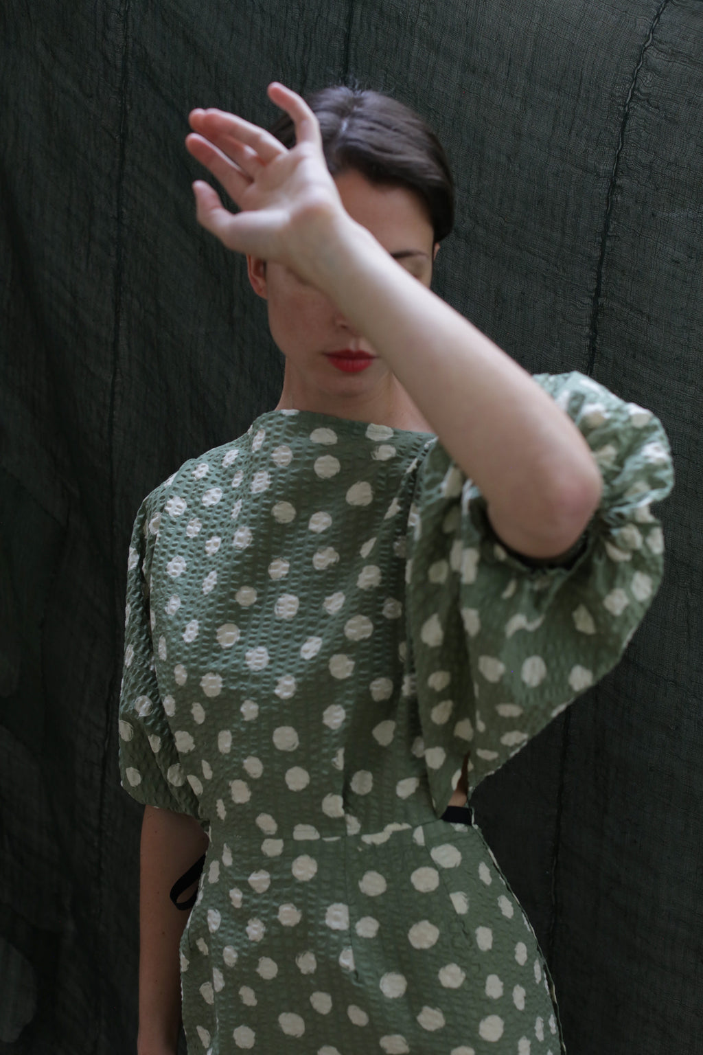 Wearing the Kimono Dress | Polkadot Seersucker Moss, a person stands before a dark fabric background, raising one arm to partially hide their face so that only their lips and part of their face can be seen.