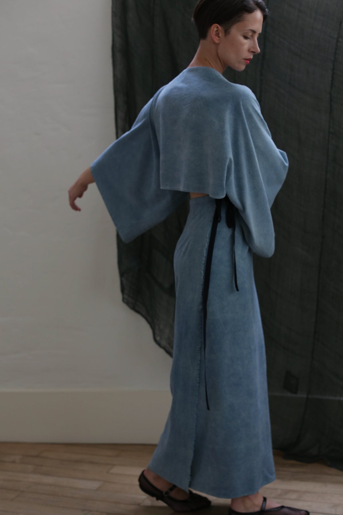 A person wearing the Kimono Dress | Silk Noil Ai Dye in light blue stands sideways with one arm extended, set against a white wall and a dark, sheer curtain.