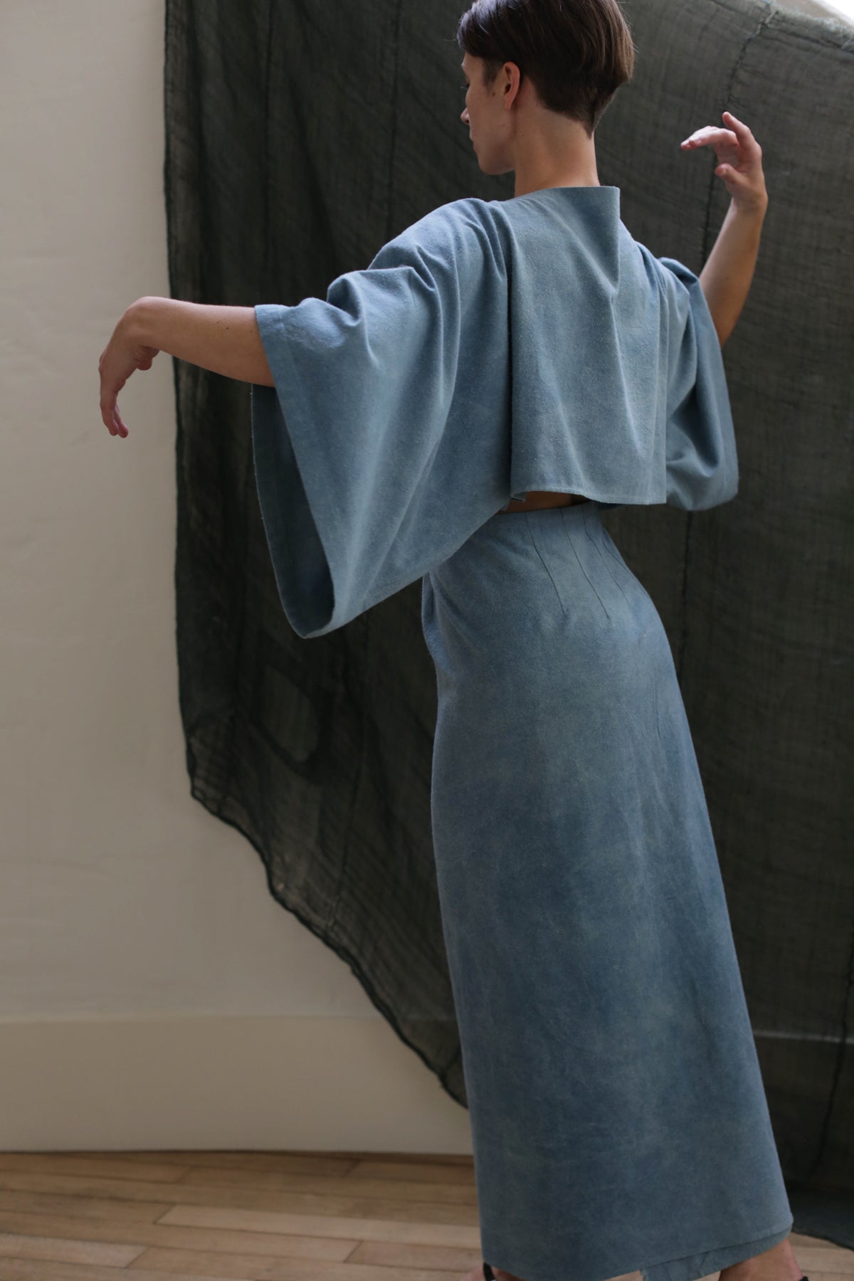 A person in the Kimono Dress | Silk Noil Ai Dye, a loose light blue kimono-inspired dress with wide sleeves, stands gracefully with arms raised, facing away from the camera before a dark semi-transparent fabric backdrop.