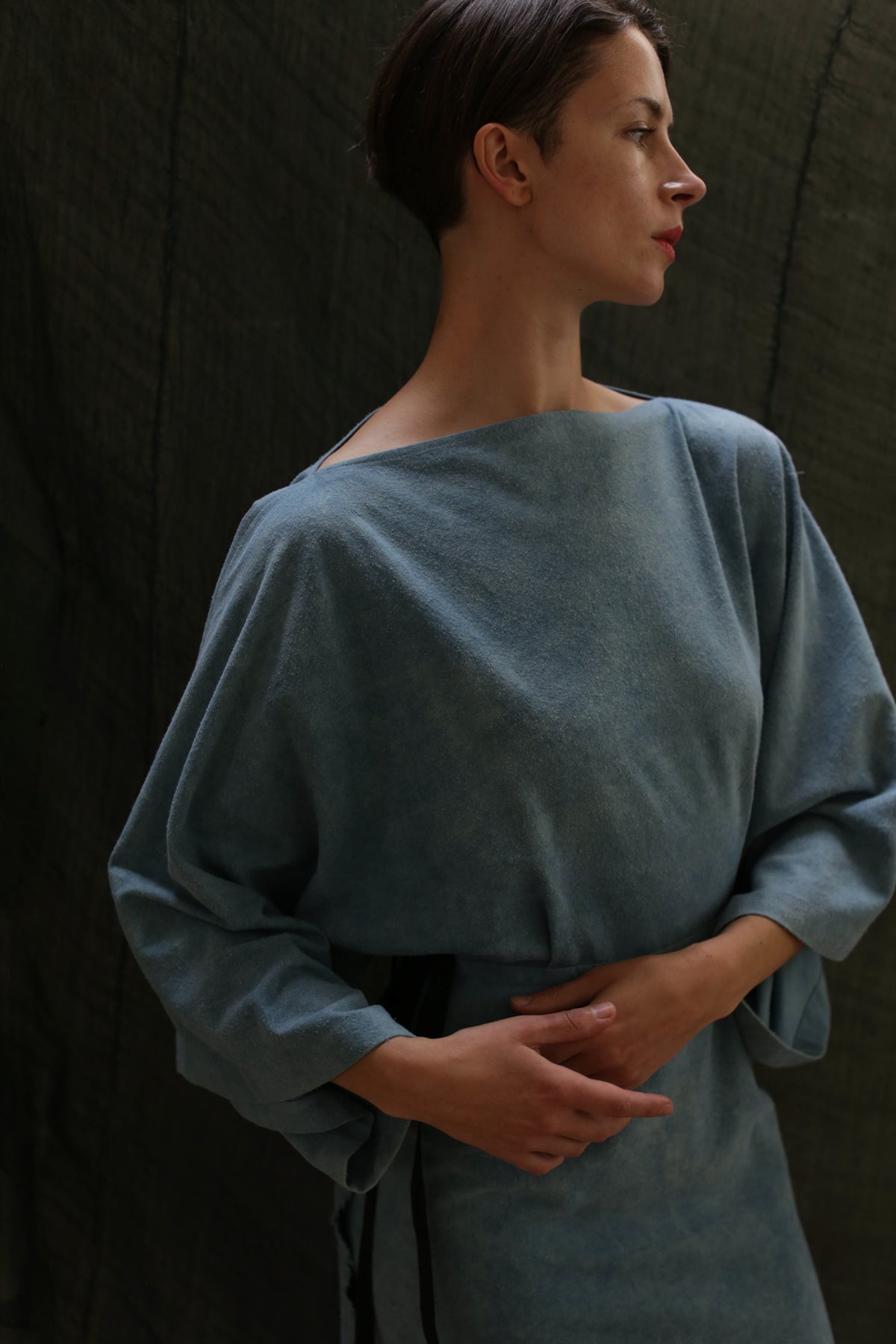 A person wearing the Kimono Dress | Silk Noil Ai Dye, a loose blue-gray long-sleeved piece crafted from soft silk noil for an elegant, textured look, stands against a dark background with hands gently clasped at the waist.