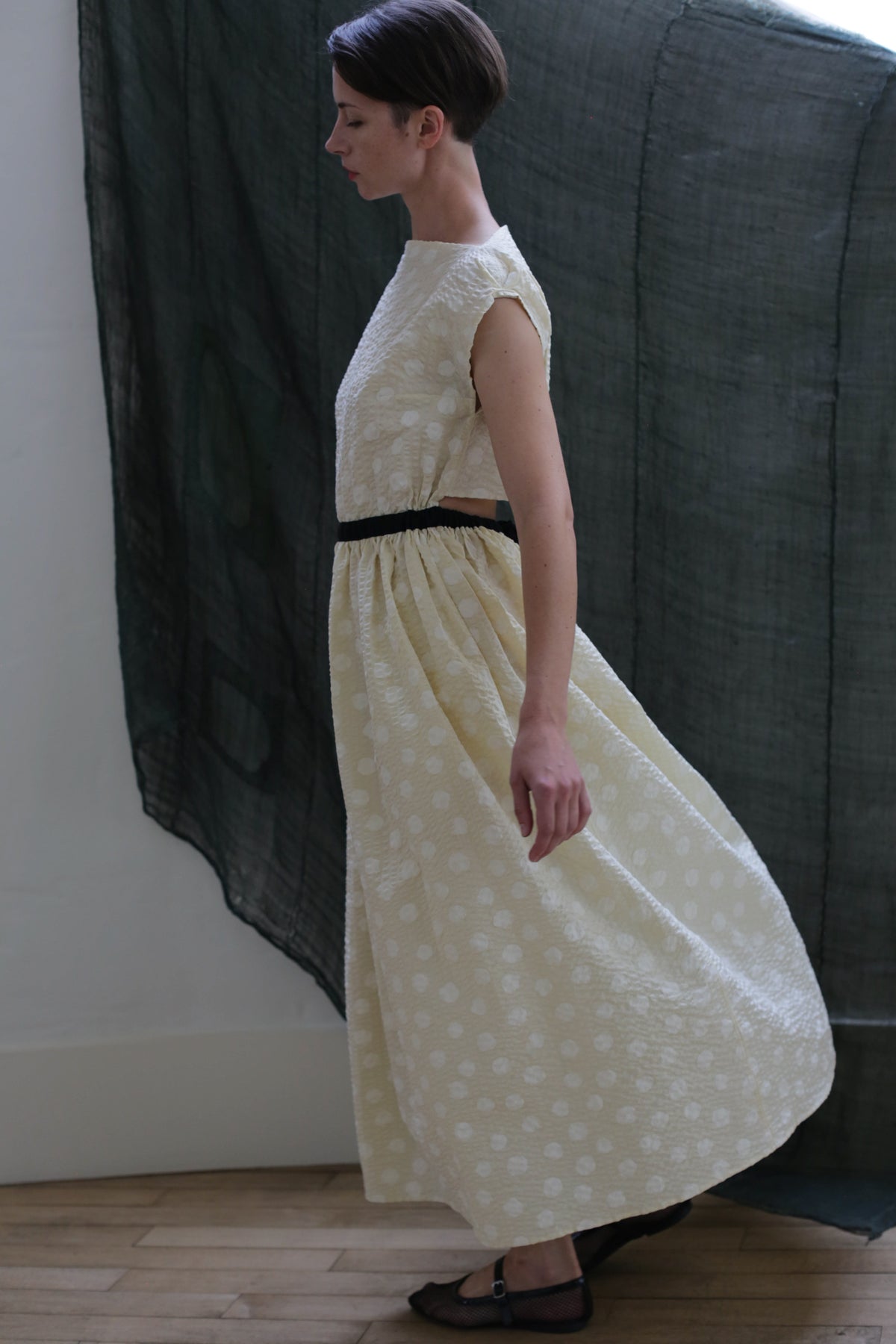 A person in the Magnolia Dress | Polkadot Seersucker Butter, a light-colored, sleeveless dress with a subtle polkadot pattern and black belt, stands in profile as the dress billows against a dark, sheer backdrop.