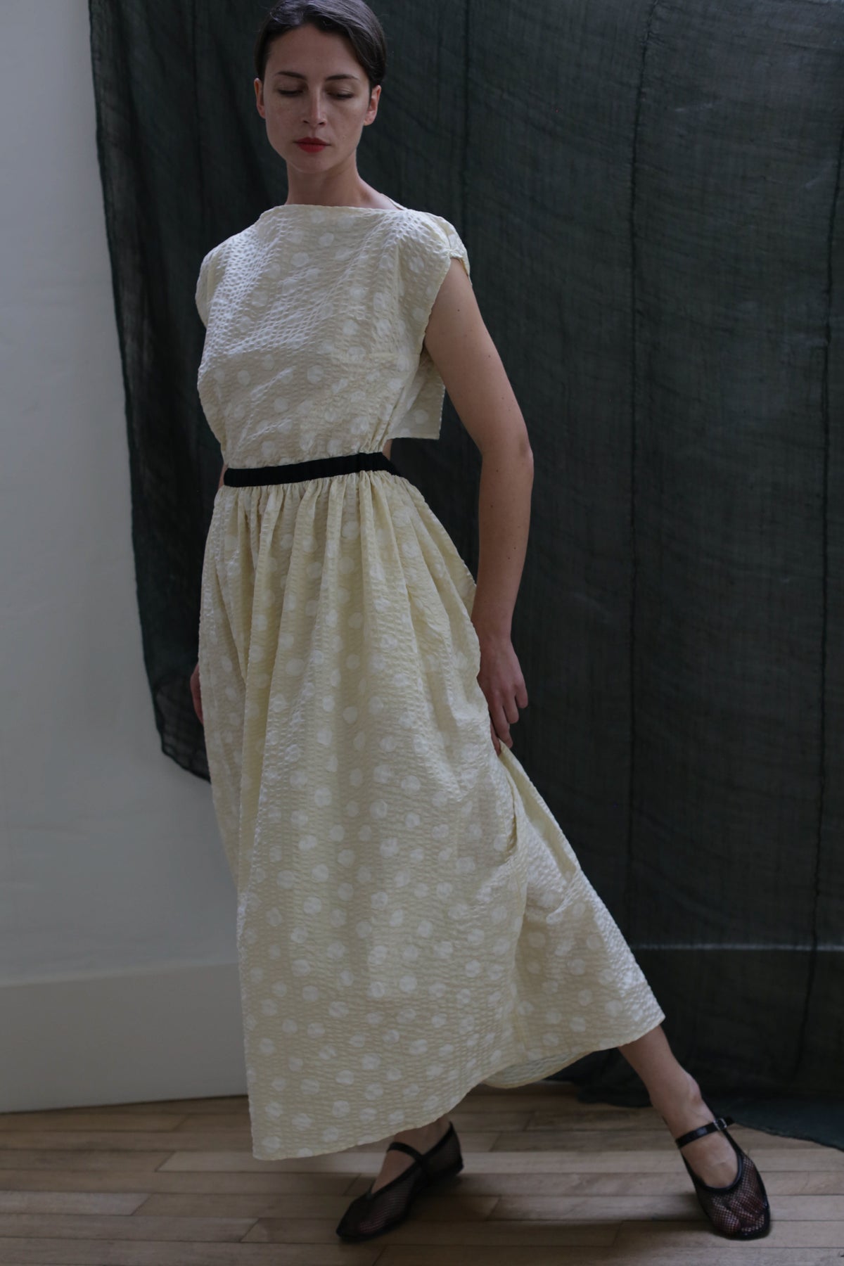 A woman wears the Magnolia Dress | Polkadot Seersucker Butter, a long cream dress with polkadots and a black belt, standing indoors with one arm bent. The background displays dark and light fabric.