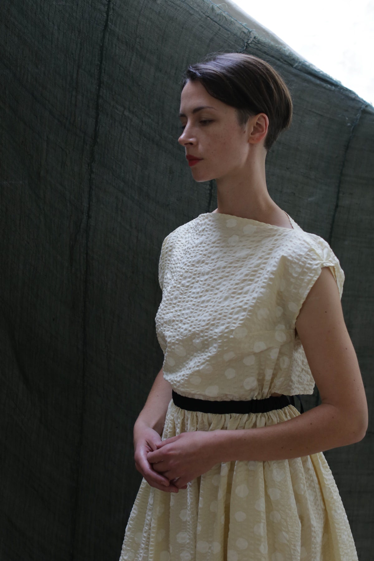 A woman with short brown hair wears the Magnolia Dress | Polkadot Seersucker Butter, a cream-colored dress with a subtle polkadot pattern and black belt, standing before a dark green fabric backdrop, looking down with hands gently clasped.