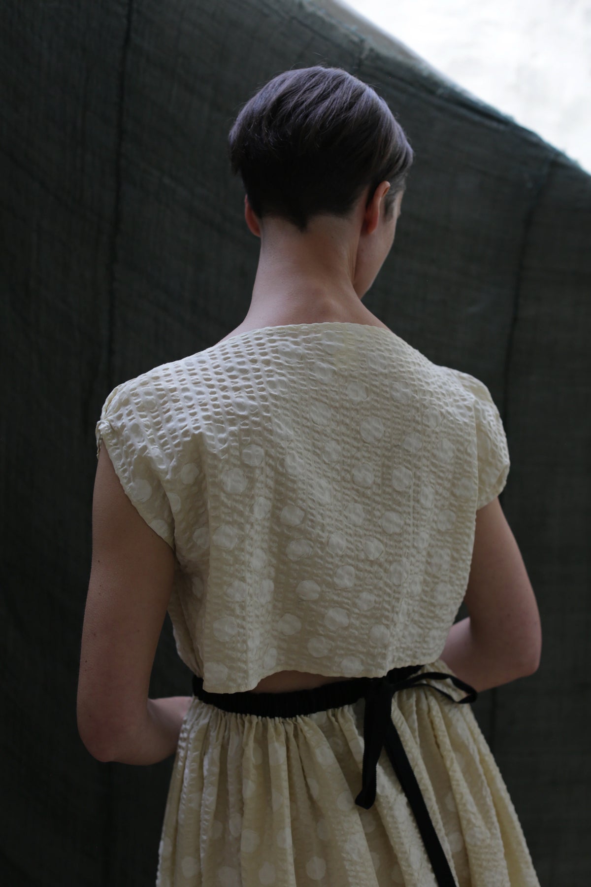 A person with short brown hair is seen from behind, wearing the Magnolia Dress | Polkadot Seersucker Butter with short sleeves and a black waist ribbon, standing before a dark fabric backdrop.