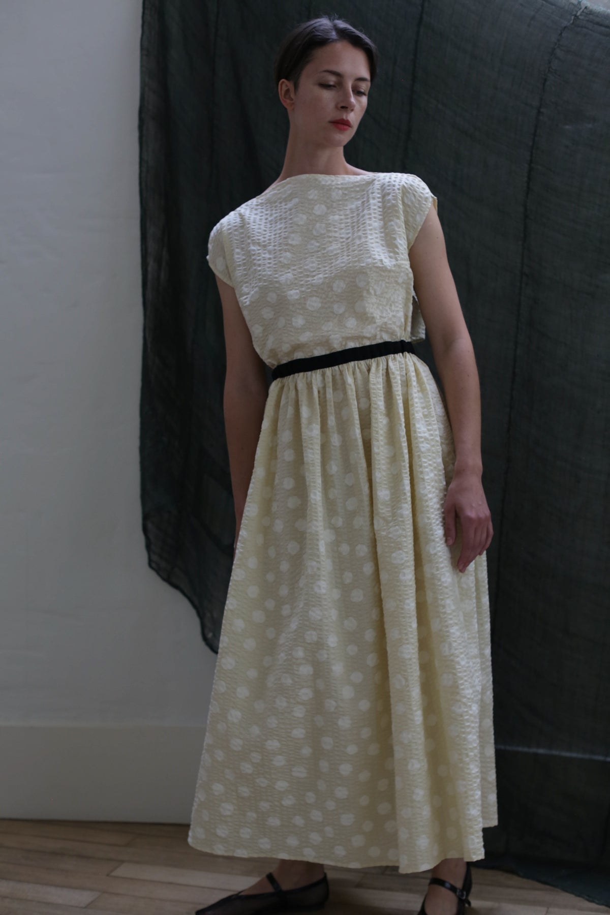 A woman stands indoors against a dark fabric backdrop, looking slightly to the side, wearing the Magnolia Dress | Polkadot Seersucker Butter—an ankle-length, light polkadot dress with a black belt.