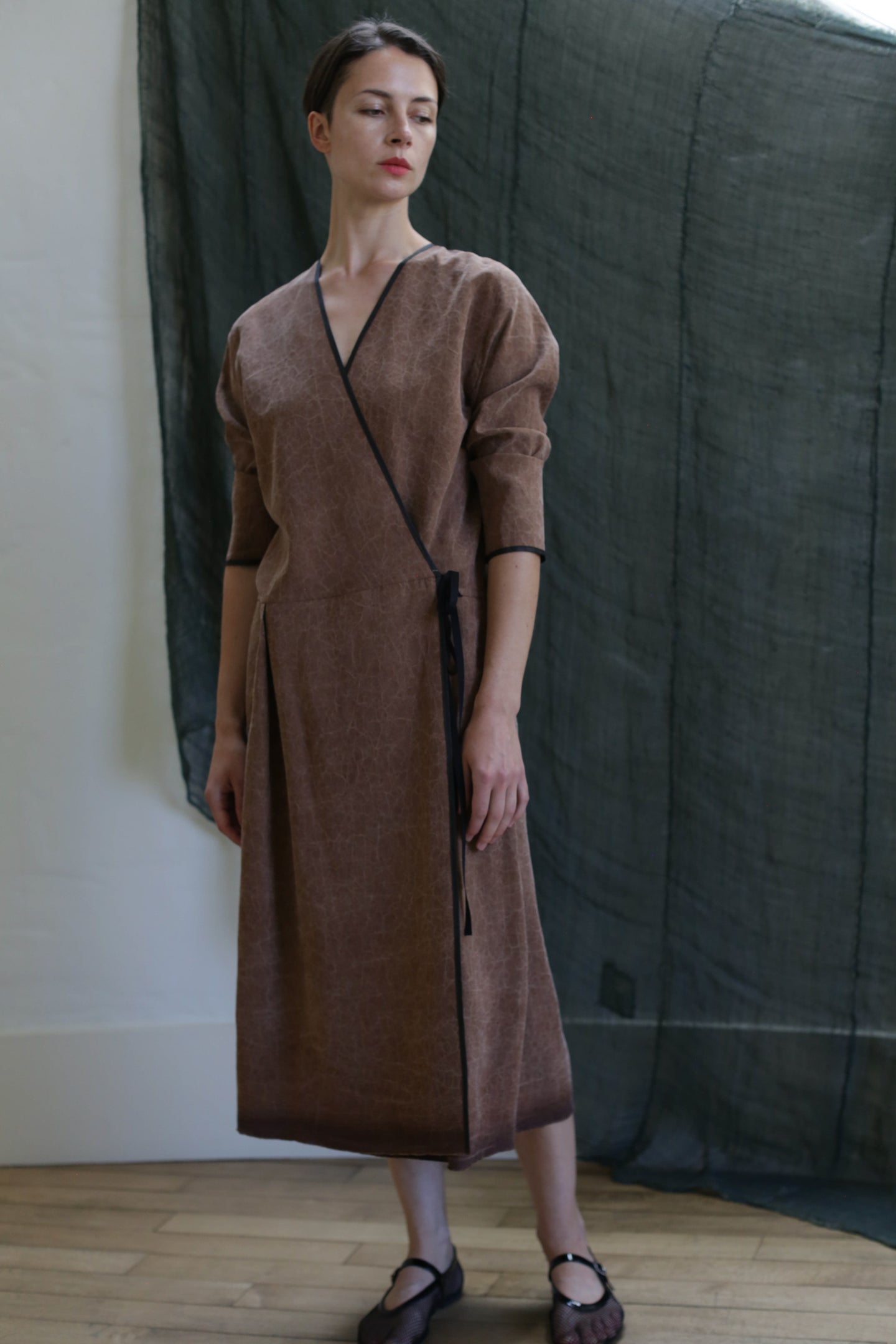 A woman stands indoors wearing the Reversible Wrap Dress with Sleeves in Xiang Yun Silk Brown, featuring three-quarter sleeves. Her hair is pulled back and she gazes slightly to the side, with dark, semi-sheer fabric in the background.