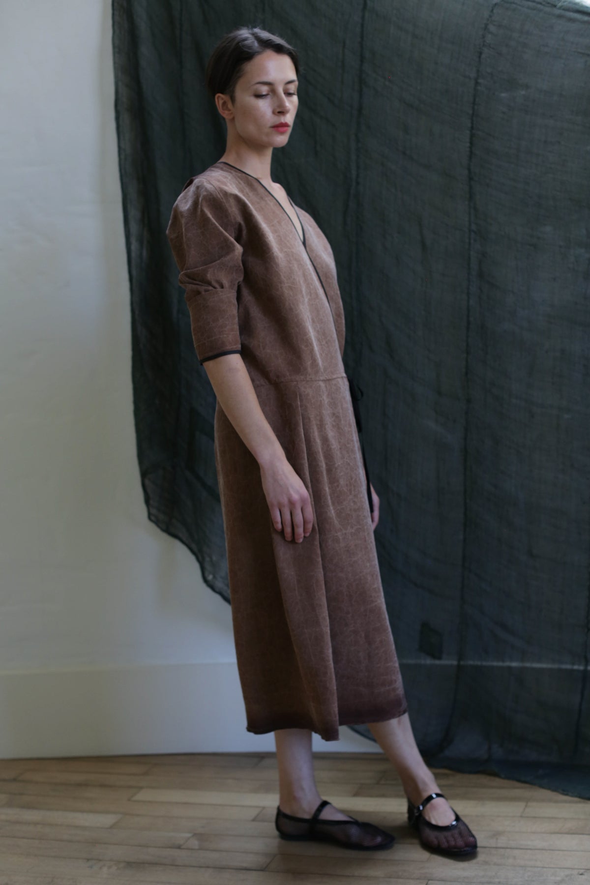 A woman with short hair stands indoors on a wooden floor, wearing black flats and the Reversible Wrap Dress with Sleeves in Xiang Yun Silk Brown. She faces sideways, with a dark sheer curtain hanging behind her.
