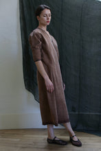 Load image into Gallery viewer, A woman with short hair stands indoors on a wooden floor, wearing black flats and the Reversible Wrap Dress with Sleeves in Xiang Yun Silk Brown. She faces sideways, with a dark sheer curtain hanging behind her.