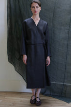 Load image into Gallery viewer, A woman stands indoors on a wooden floor, wearing the Reversible Wrap Dress with Sleeves in Xiang Yun Silk Brown. The ankle-length dress features long sleeves and a subtle sheen. A sheer dark fabric hangs as a backdrop behind her.