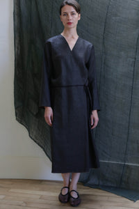 A woman stands indoors on a wooden floor, wearing the Reversible Wrap Dress with Sleeves in Xiang Yun Silk Brown. The ankle-length dress features long sleeves and a subtle sheen. A sheer dark fabric hangs as a backdrop behind her.