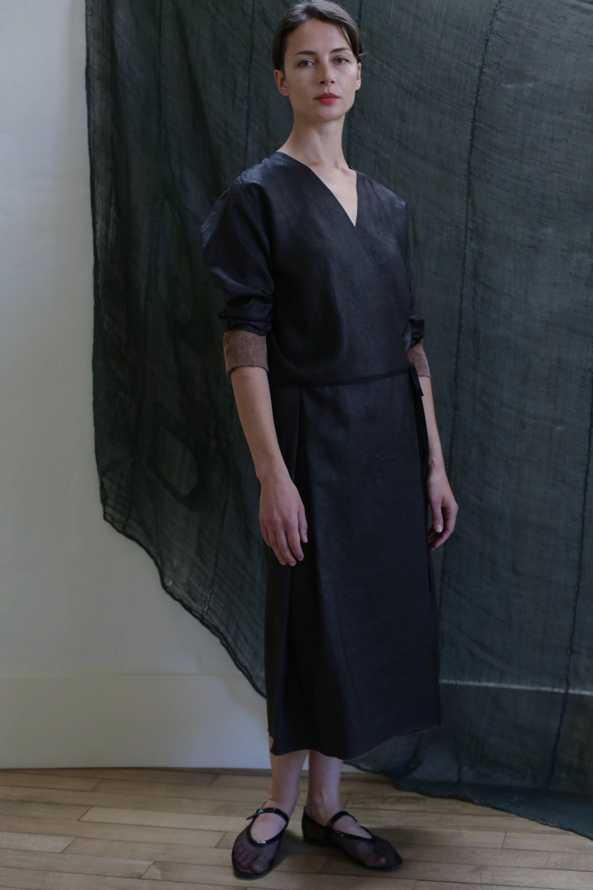A woman stands indoors in a long-sleeved Reversible Wrap Dress with Sleeves | Xiang Yun Silk Brown and black shoes, framed by a sheer dark green fabric on the wall in a minimalistic setting.