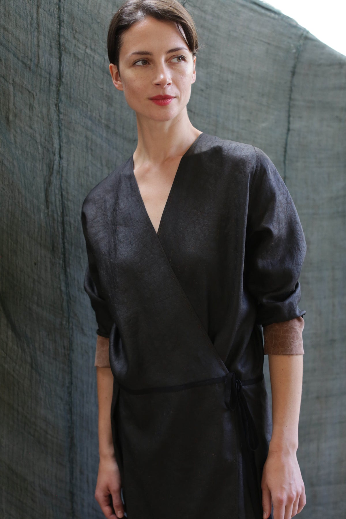 A woman with short brown hair stands before a textured gray backdrop, wearing the Reversible Wrap Dress with Sleeves | Xiang Yun Silk Brown—a dark, long-sleeved wrap dress featuring light brown cuffs. She gazes slightly to her left with a natural expression.
